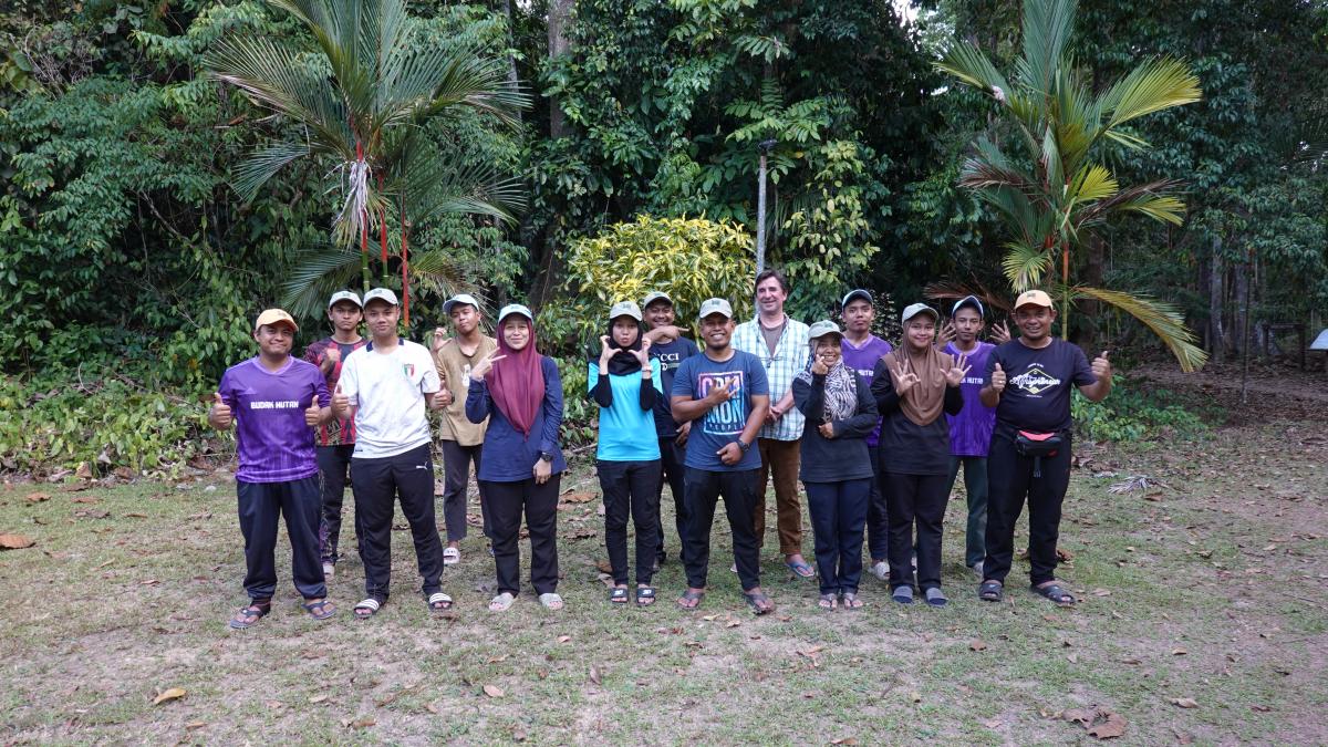 a group of people (the census team) stand in front of the edge of the forest, smiling. Many are holding up peace signs or thumbs up. 