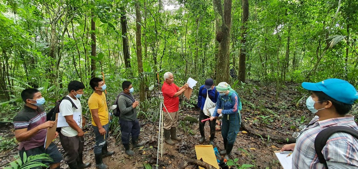 Technicians stand in a tropical forest, their focus on a man in a red shirt.