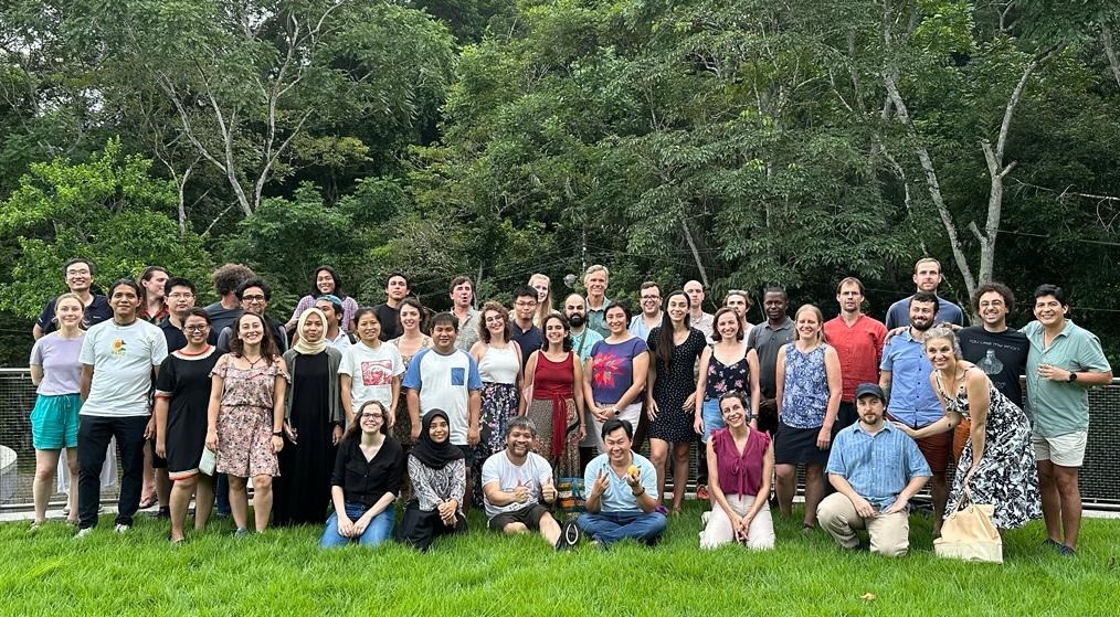 A large group of people standing and sitting on grass with forest in the background, all smiling at the camera.