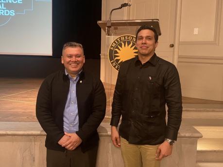Milton Solano and David Mitre stand in front of a stage, smiling at the camera. 