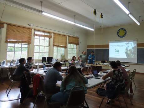 Workshop participants sit at a u-shaped table facing a presentation in a classroom with large windows