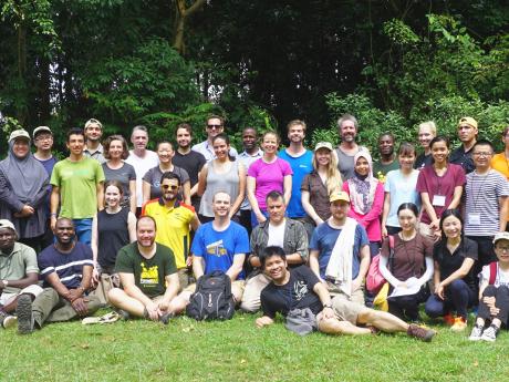 A group photo of the 53 workshop participants, assembled in 2 rows, some standing, others sitting, in front of cluster of trees.