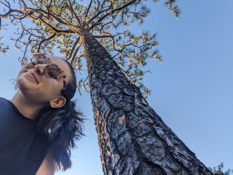 A woman in sunglasses smiles in a selfie with a tall tree, blue sky in background